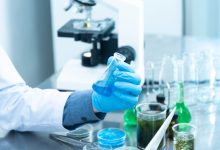 Cientista usando luvas analisa um líquido azul em um laboratório, com microscópio e vidrarias ao fundo.