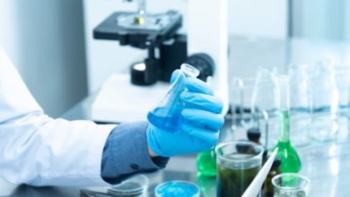 Cientista usando luvas analisa um líquido azul em um laboratório, com microscópio e vidrarias ao fundo.