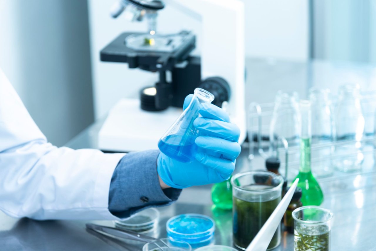 Cientista usando luvas analisa um líquido azul em um laboratório, com microscópio e vidrarias ao fundo.