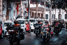 Motociclistas circulam por uma movimentada rua urbana ao lado de táxis e outros veículos.