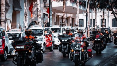 Motociclistas circulam por uma movimentada rua urbana ao lado de táxis e outros veículos.