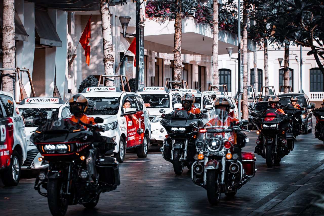 Motociclistas circulam por uma movimentada rua urbana ao lado de táxis e outros veículos.